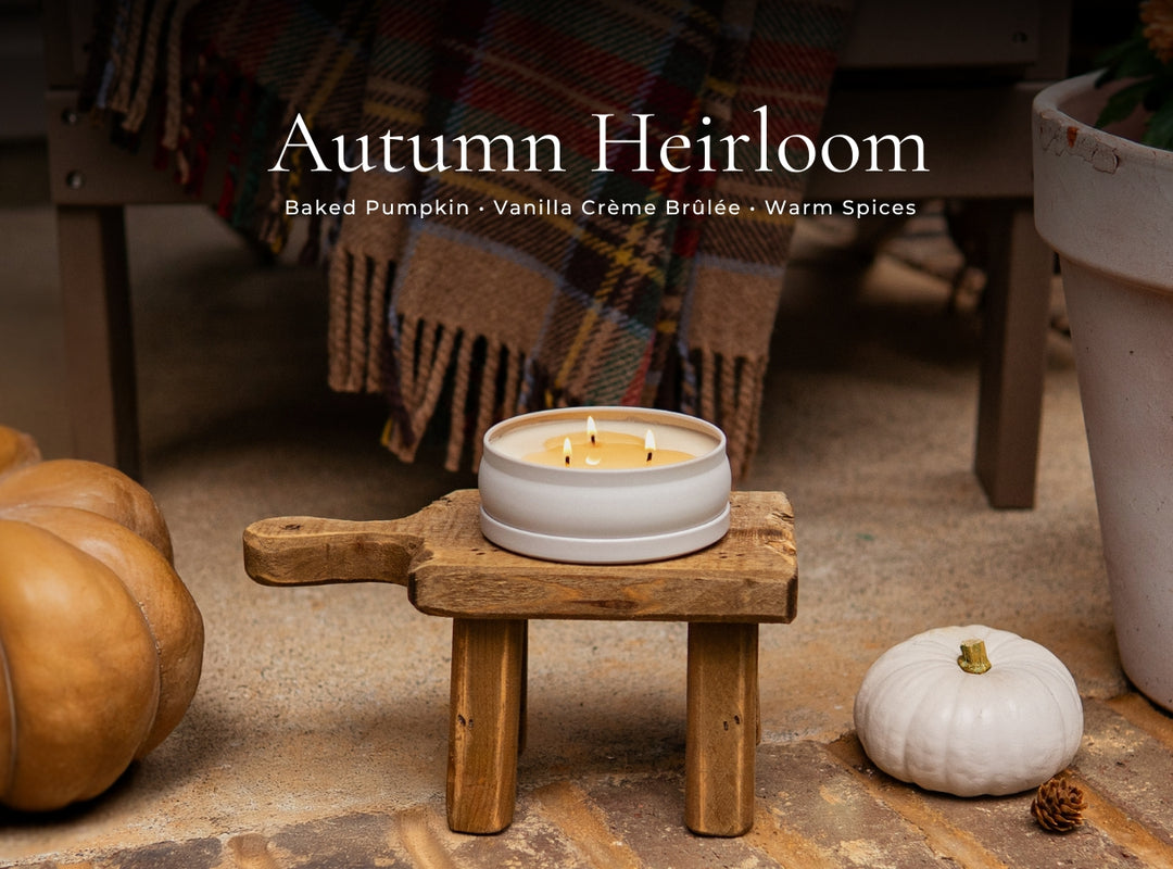 A lit candle in a white ceramic holder sits on a small wooden stool in a styled autumn setting with pumpkins, a plaid blanket, and the text 'Autumn Heirloom' at the top.
