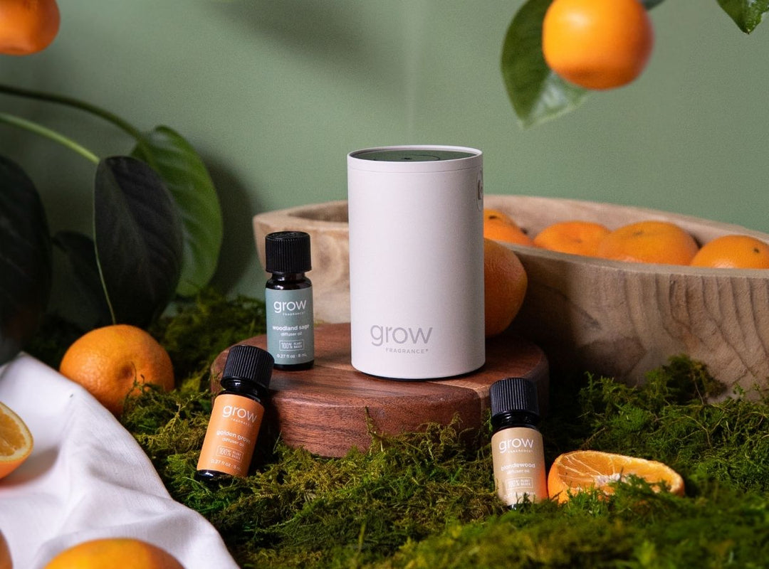 Grow Fragrance waterless diffuser in matte white sitting on a wooden pedestal, surrounded by three diffuser oil bottles — Woodland Sage, Golden Grove, and Blondewood — arranged on a bed of green moss with fresh oranges, citrus slices, and a wooden bowl of oranges against a sage green backdrop. (featured)