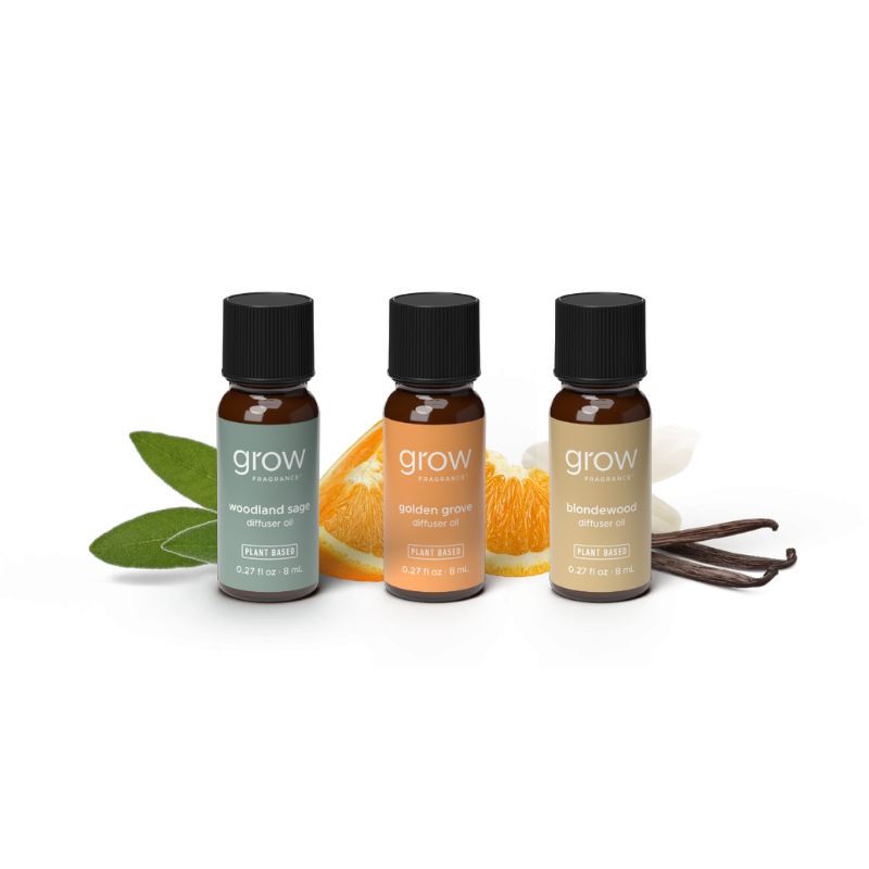 Three small amber glass essential oil bottles with black caps from grow naturally, arranged on a white background with flavor props including green leaves, orange slices, white blossoms, and vanilla beans. 