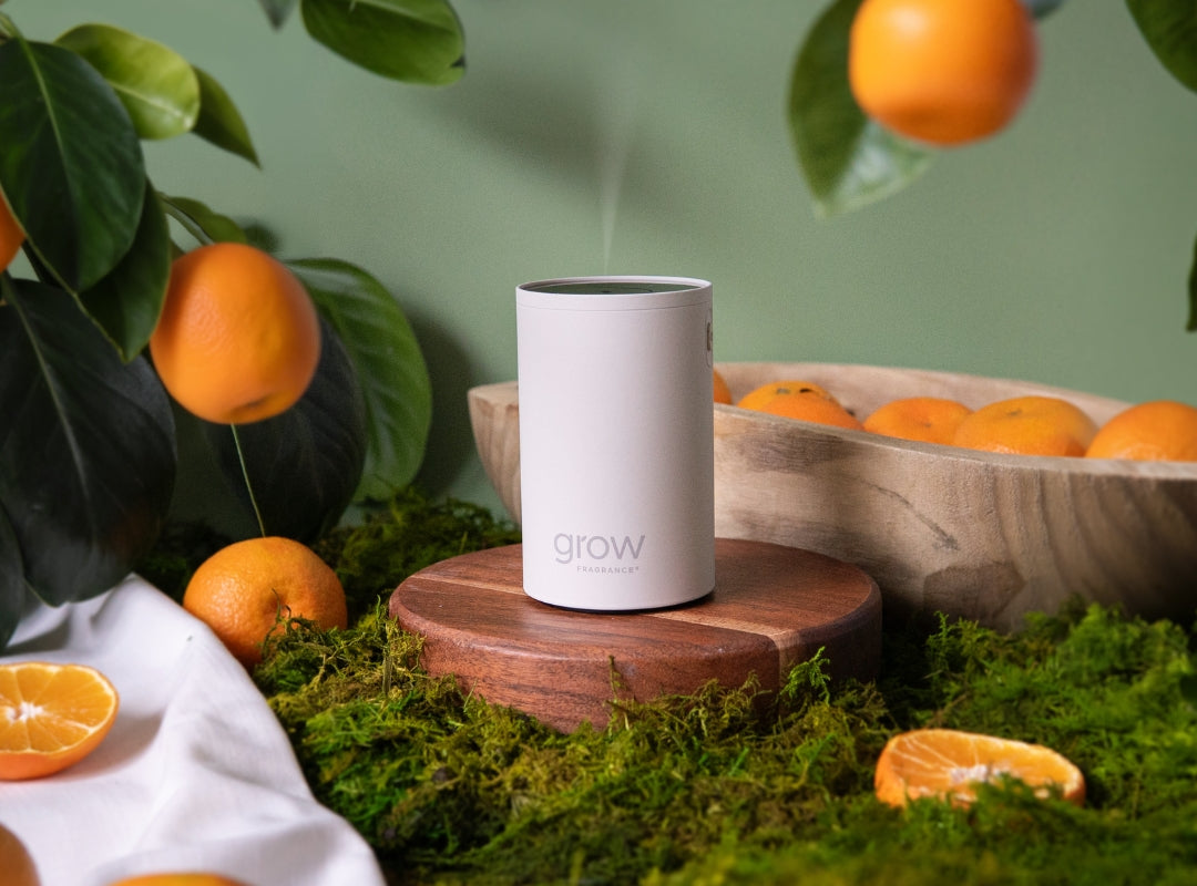 A white cylindrical Grow diffuser sits on a round wooden pedestal, surrounded by green moss, oranges, and a wooden bowl, with a soft green background.