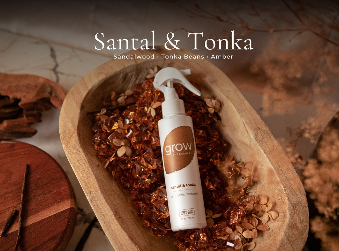A bottle of Grow Hair Growth Serum in Santal & Tonka scent, placed in a wooden bowl filled with sandalwood pieces and dried botanicals. The background is a warm, rustic setting with a marble surface and dried flowers, and the text 'Santal & Tonka' is displayed at the top.