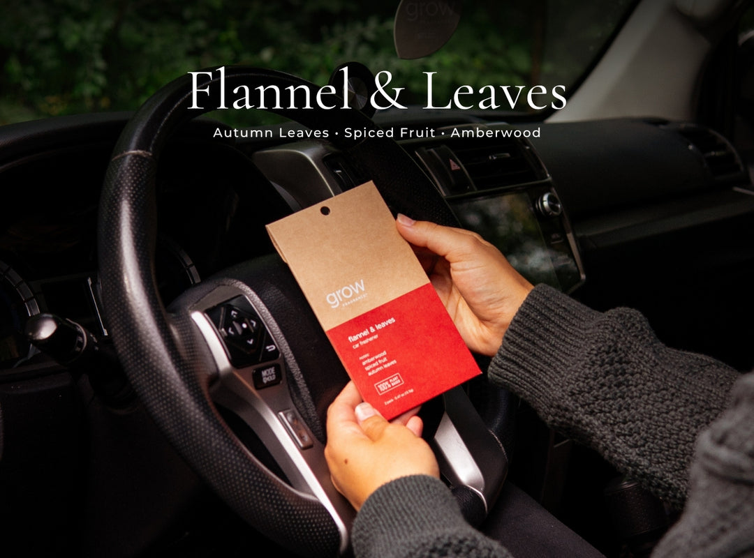 A person inside a car holds a packaged car fragrance tag labeled 'Flannel & Leaves' by Grow. The tag is kraft brown on the top half and red on the bottom half.
