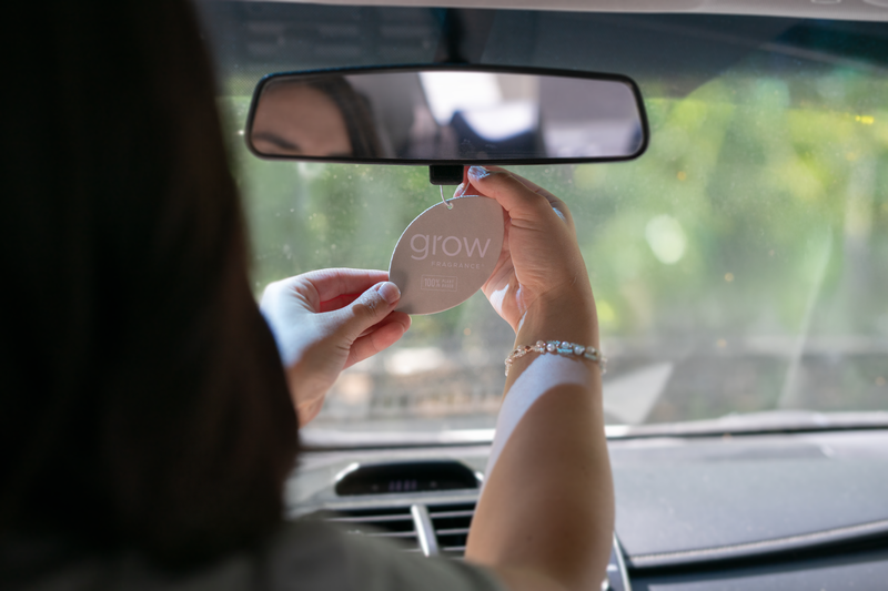 Person holding a 'grow' branded car air freshener in front of a car mirror.