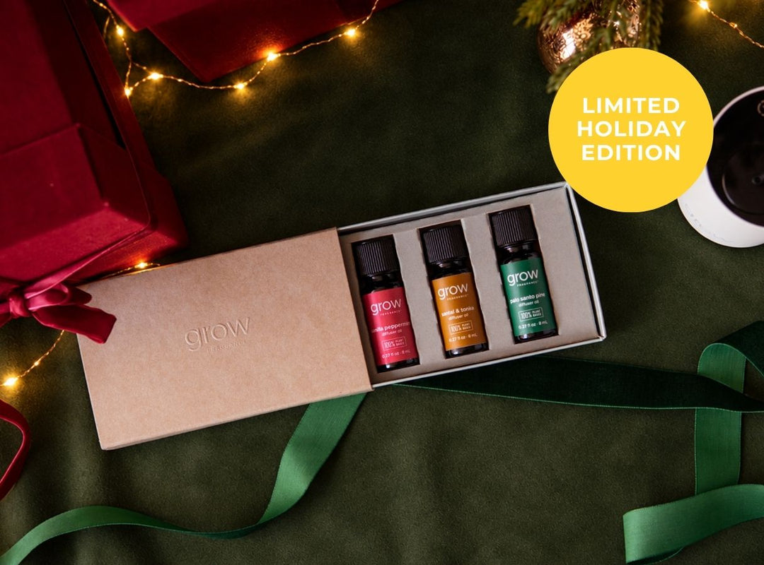 A boxed set of three GLOW essential oils displayed in an open kraft gift box on a dark green fabric surface, with red gift boxes, green ribbons, warm string lights, and a yellow 'LIMITED HOLIDAY EDITION' badge. (featured)