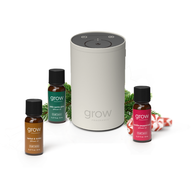 A Grow Fragrance diffuser set on a white background, featuring a central light gray cylindrical diffuser surrounded by three small fragrance bottles: amber, green, and red, with pine branches and candy canes as styling props. (featured)