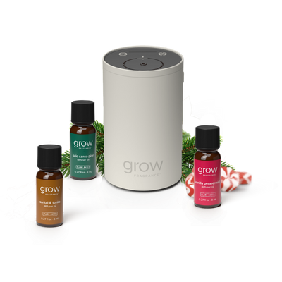 A Grow Fragrance diffuser set on a white background, featuring a central light gray cylindrical diffuser surrounded by three small fragrance bottles: amber, green, and red, with pine branches and candy canes as styling props. (featured)