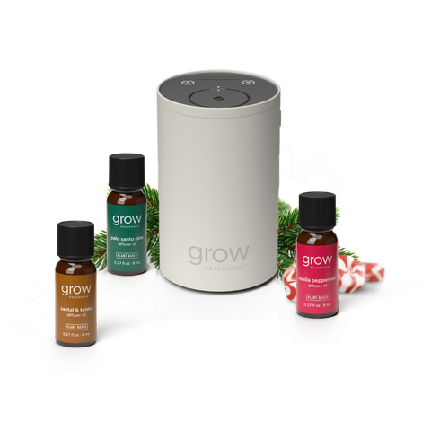 A Grow Fragrance diffuser set on a white background, featuring a central light gray cylindrical diffuser surrounded by three small fragrance bottles: amber, green, and red, with pine branches and candy canes as styling props. (featured)