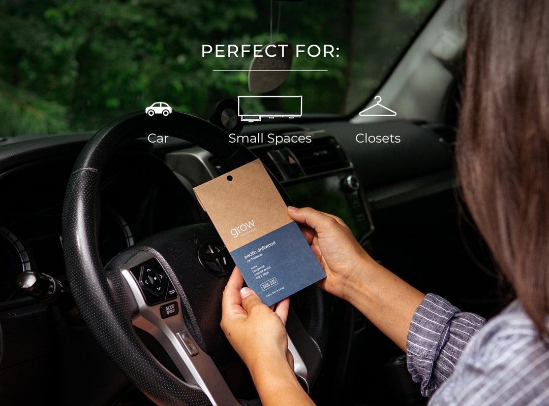Person holding a small blue and brown package labeled Pacific Driftwood car freshener inside a car, with text indicating it's perfect for car, small spaces, and closets.