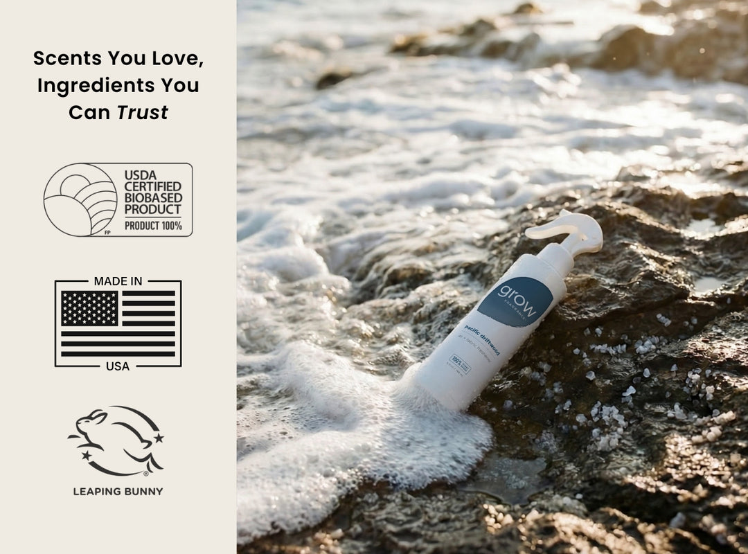 Pacific Driftwood bottle on a rocky beach with branding and certification logos. Scents You Love, Ingredients You Can Trust / USDA Certified Biobased Product 100% / Made in USA / Leaping Bunny 