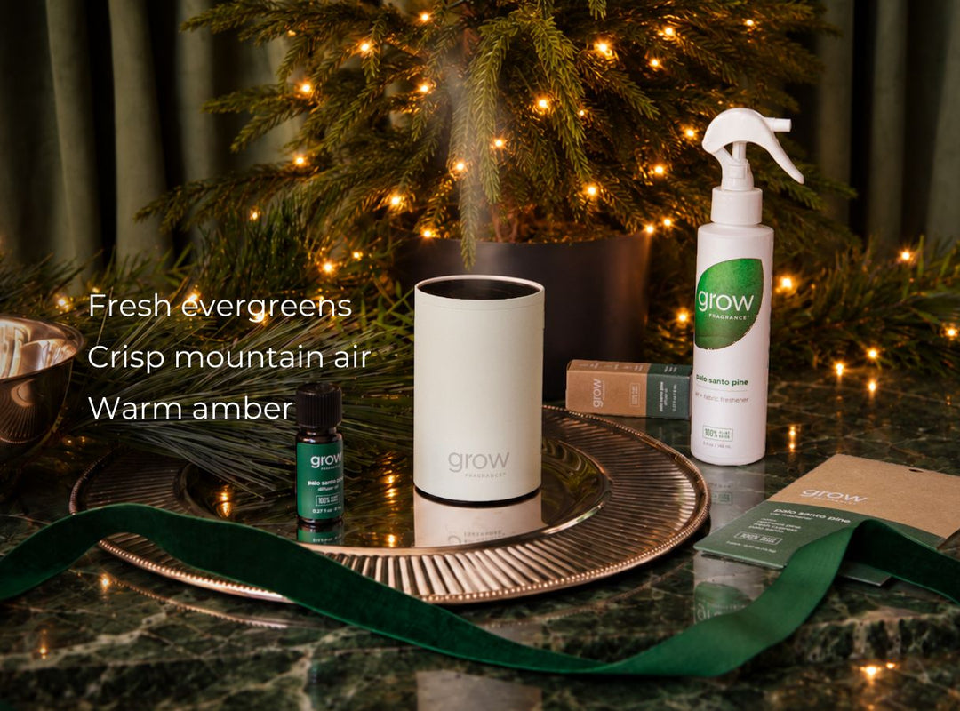 A white cylindrical Grow brand diffuser emitting a mist, displayed on a round metallic tray next to a white Grow spray bottle and a small green essential oil bottle, set on a dark marble surface with a Christmas tree and warm string lights in the background. (featured)