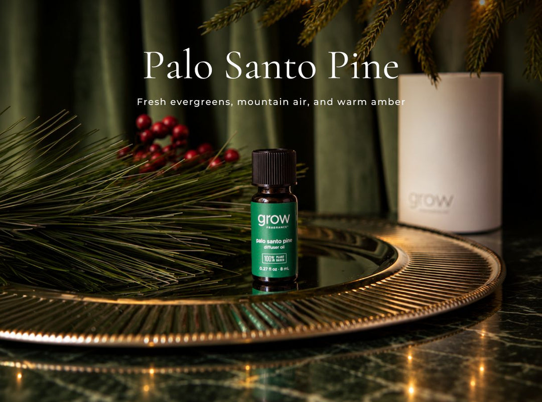 A small dark green bottle labeled 'grow' and 'Palo Santo Pine' sits on a round gold tray, styled with pine branches, red berries, and a white cylindrical container against a dark green curtain background. (featured)