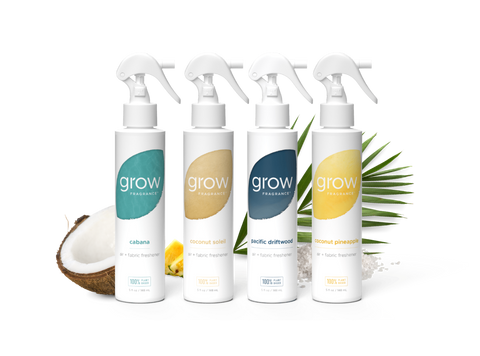 Four white spray bottles from the brand grow professionally lined up on a white background, styled with a halved coconut, lemons, and green palm leaves. The bottles feature colorful labels in teal, beige, navy, and yellow, representing the flavors: cabana, coconut sole, Pacific Delitt, and Coconut Pine森.