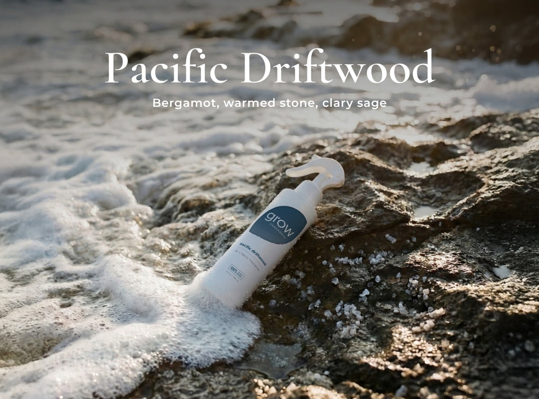 A white spray bottle labeled 'grow' lies on wet rocks surrounded by foamy surf, with overlaid text reading 'Pacific Driftwood' and 'Bergamot, warmed stone, clary sage'.