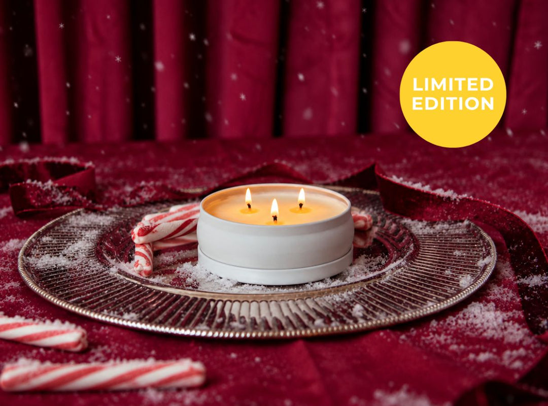 A lit white candle in a round tin sits on a decorative silver tray, surrounded by candy canes and white snow-like dust, against a deep red fabric backdrop with a yellow 'LIMITED EDITION' badge in the top right. (featured)