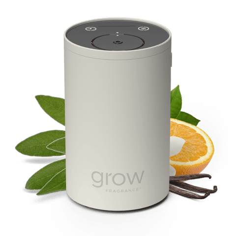 A cylindrical Grow Fragrance diffuser in a light warm gray color, styled with fresh green sage leaves, a sliced orange, and vanilla beans on a clean white background.