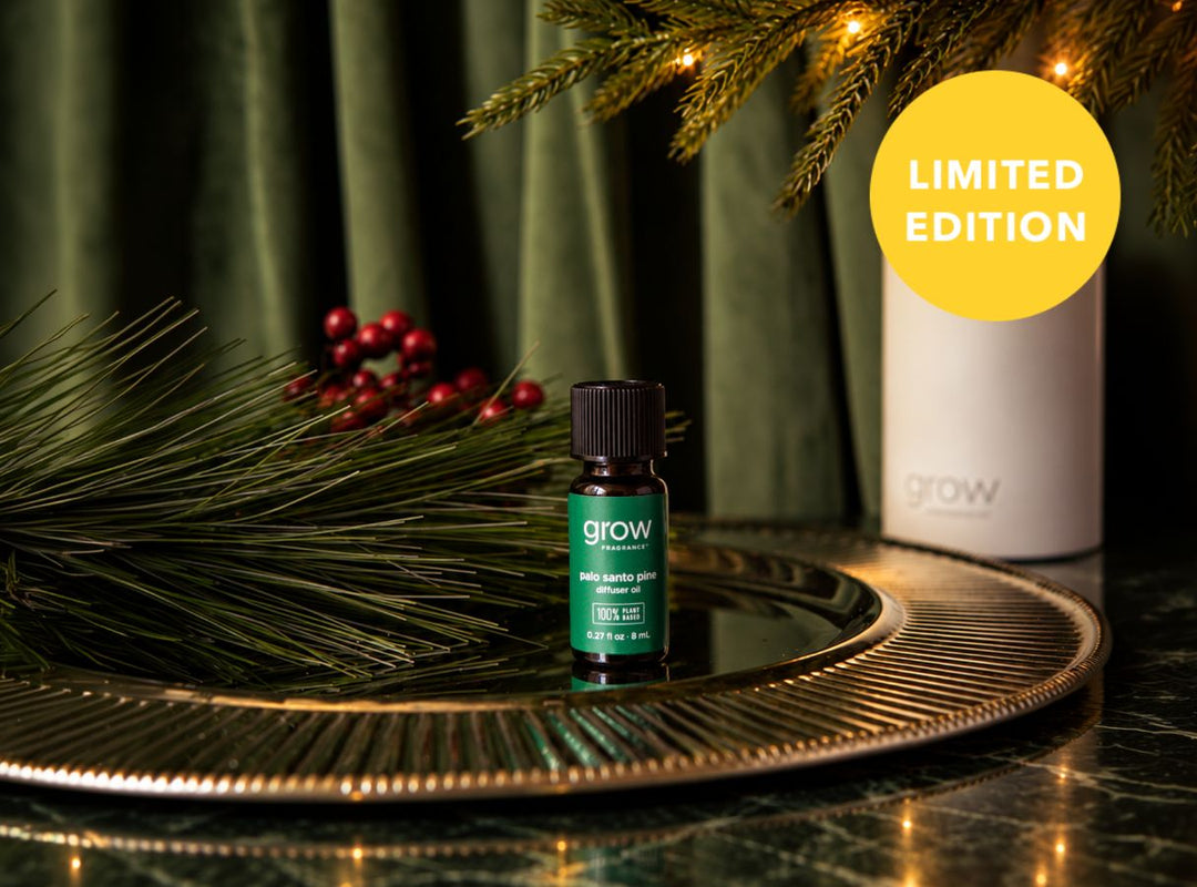 A small green and black grow essential oil bottle sits on a round mirrored tray, surrounded by pine branches, red berries, and a white cylindrical container in the background. A yellow circle in the top right reads 'LIMITED EDITION'. (featured)