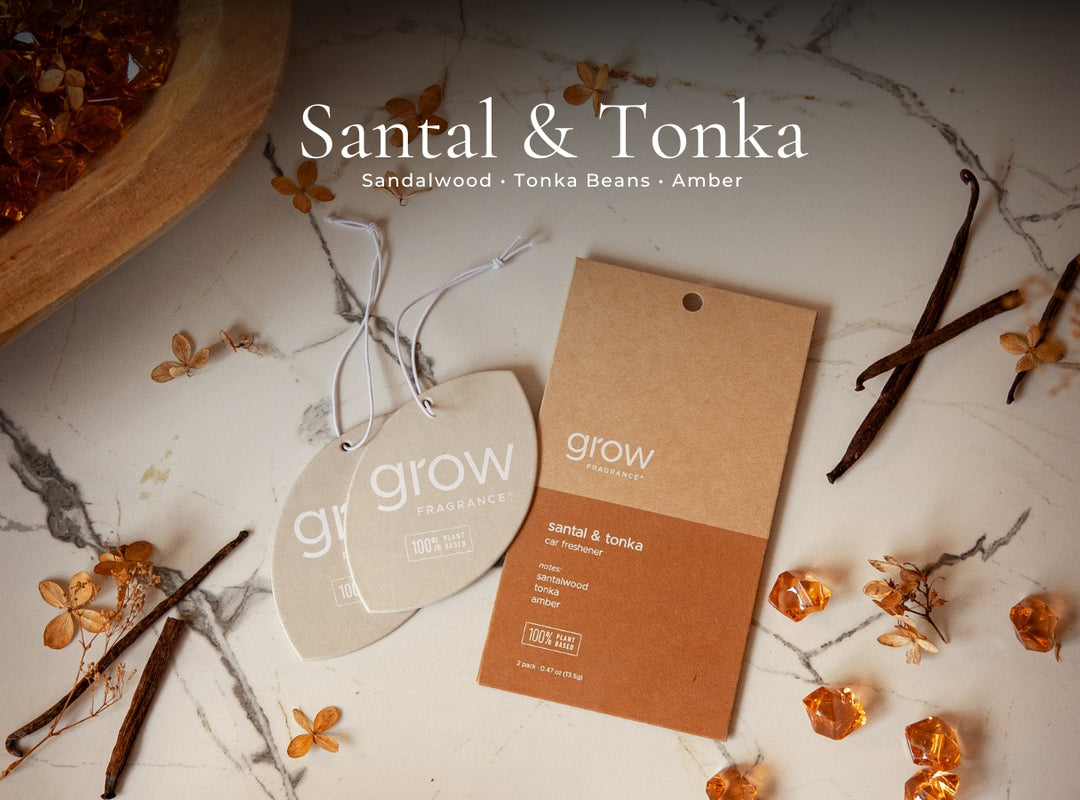 A styled flat-lay of a Grow Fragrances Santal & Tonka scent card and matching heart-shaped tag, surrounded by amber crystals, vanilla beans, and dried flowers on a white marble surface. The text 'Santal & Tonka' is displayed at the top.