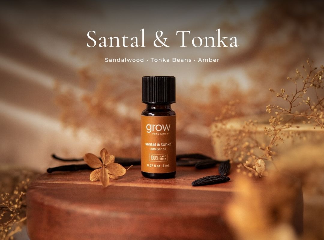 A small amber glass essential oil bottle labeled 'grow' and 'santal & tonka' stands on a round wooden pedestal with vanilla beans and dried botanicals around it. The background is warm beige and softly blurred, with the text 'Santal & Tonka' and 'Sandalwood • Tonka Beans • Amber' at the top. (featured)